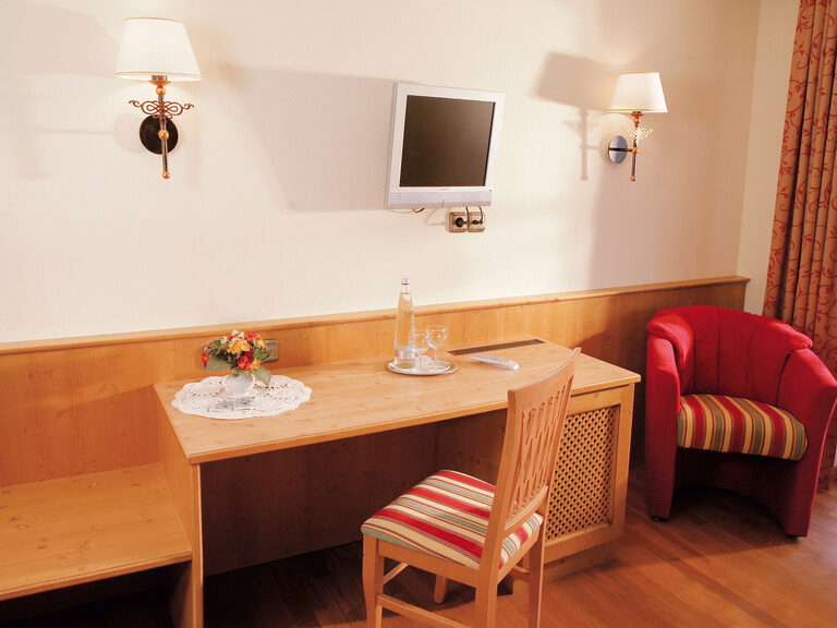 Ein Tisch mit Stuhl aus Holz sowie ein roter Sessel in einer Ecke in einem Hotelzimmer im Happinger Hof