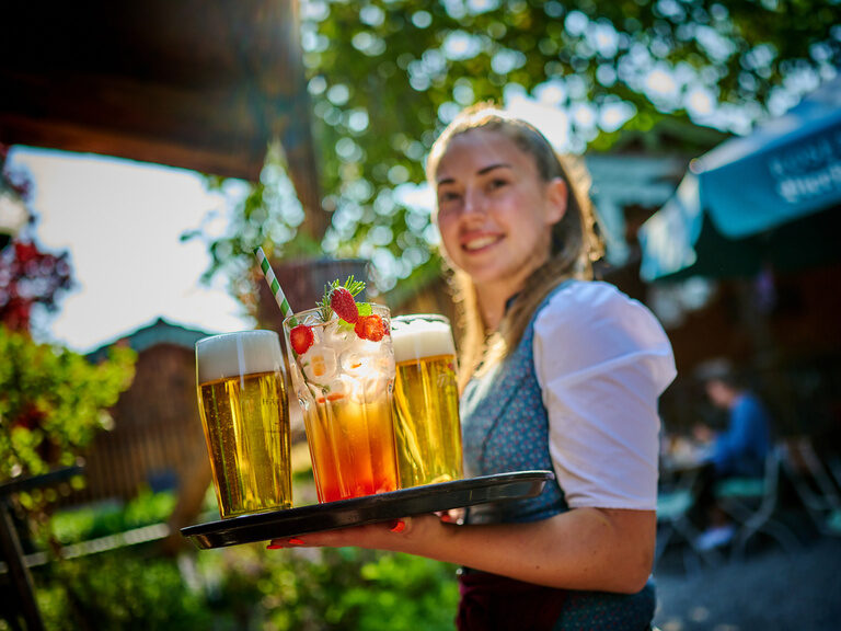 Eine gut gelaunte Bedienung trägt ein Tablet mit Getränken durch den Biergarten des Hotels Happinger Hof in Oberbayern.