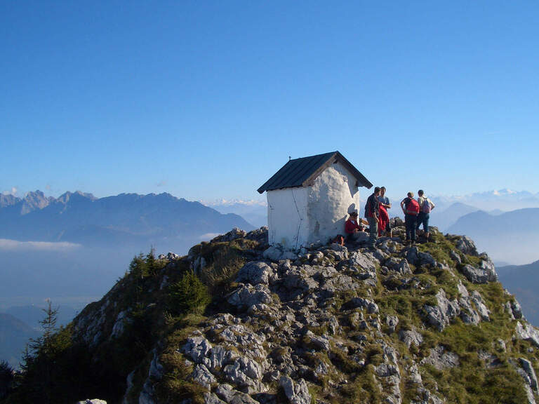 Mehrere Wanderer stehen auf einem kleinen Gipfel in den Alpen auf dem ein kleines Häuschen steht mit Ausblick auf das Umland