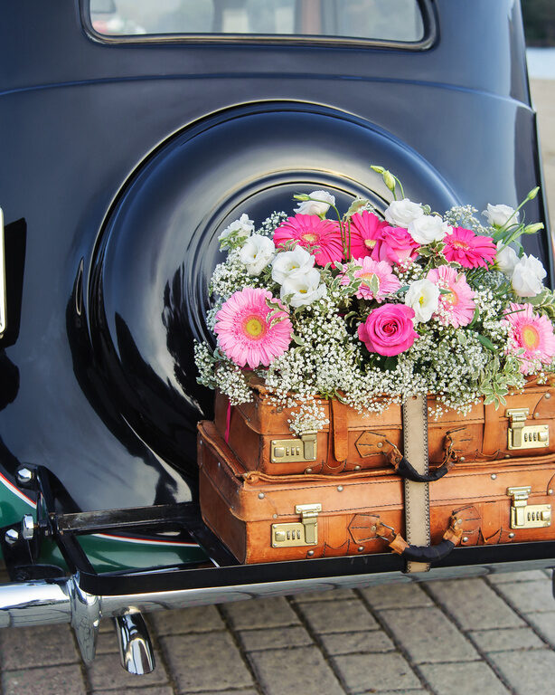 Ein schwarzer Oldtimer, der als Hochzeitsauto genutzt wird, hat zwei Koffer und Blumen am Heck
