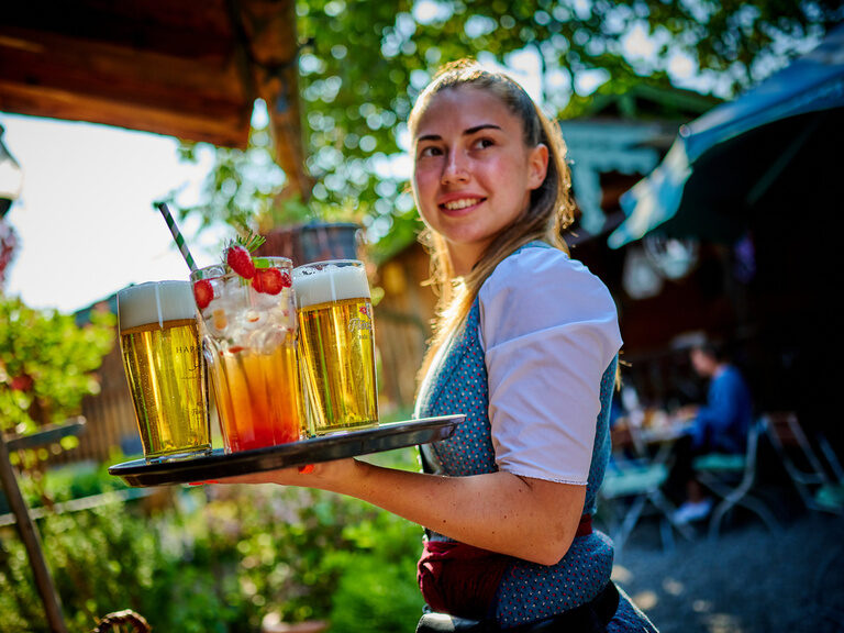 Lächelnde Bedienung trägt Tablet voller Getränke an sonnigem Tag durch Biergarten