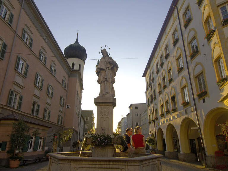 Springbrunnen aus Stein mit Kirchenfigur bei Dämmerung in oberbayerischer Altstadt