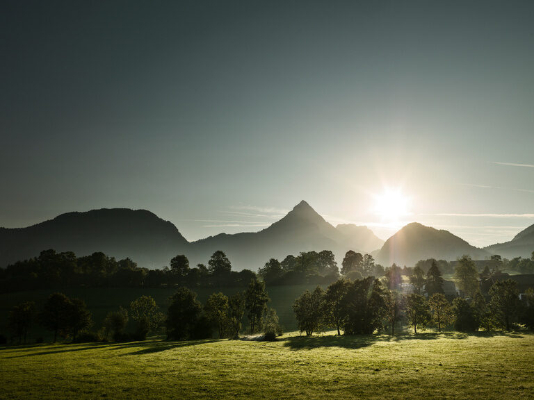 Sonne scheint zwischen Gebirge hindurch, davor grüne Wiesen und Bäume