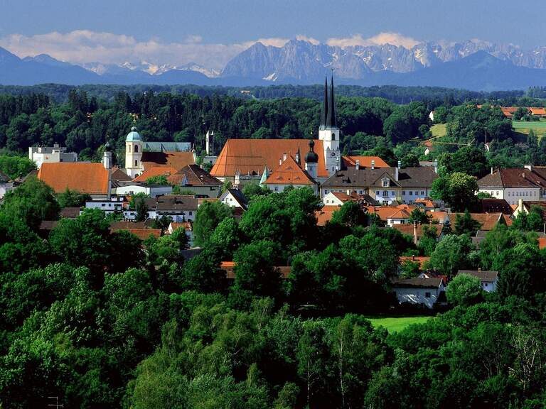 Stadt Altötting mit Kirche nd mehreren Häusern, umgeben von Wäldern, dahinter Gebirge,