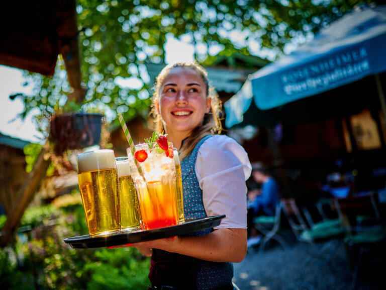 Lächelnde Bedienung trägt Tablett mit Getränken an sonnigem Tag durch den Biergarten des Hotels