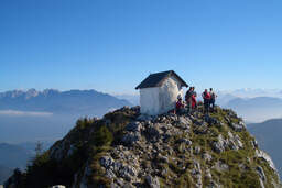 Mehrere Wanderer stehen auf einem kleinen Gipfel in den Alpen auf dem ein kleines Häuschen steht mit Ausblick auf das Umland