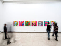 Mehrere Besucher begutachten die bunten Gemälde einer Kunstausstellung in einem weißen Raum