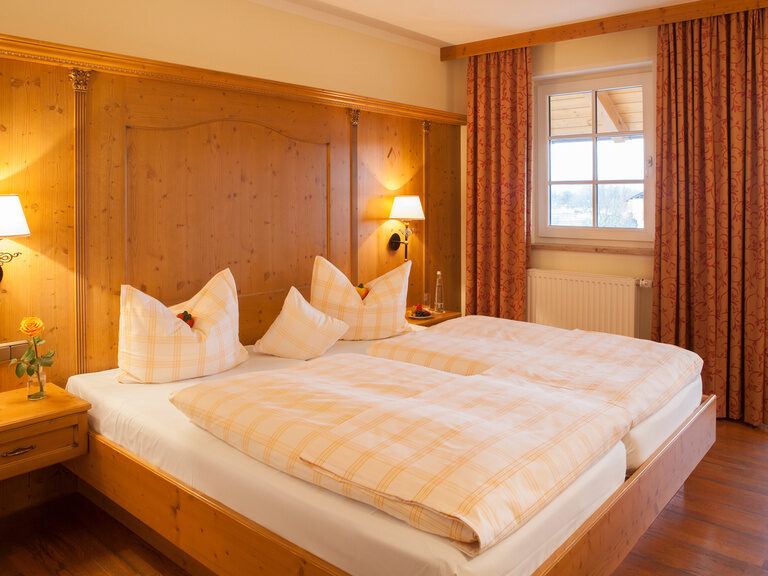 Ein Doppelbett in einem Hotelzimmer, das im Landhausstil gehalten ist mit Holzmöbeln und zwei Fenster, sowie gemütlicher Beleuchtung