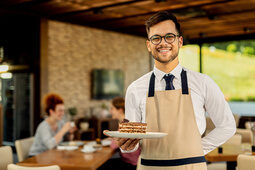 Ein junger Mann der als Biergarten-Aushilfe arbeitet, hält ein einen Teller mit einem Kuchen
