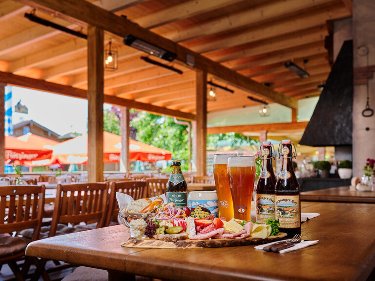 Brotzeitplatte auf Tisch in Biergarten, mit Weizengläsern, Brotkorb, Aufstrichen, Käse und Wurst