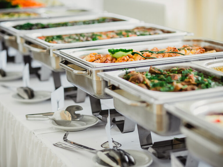 Aufgereihte Catering-Speisen auf einem Tisch mit weißer Tischdecke und silbernen Wärmebehältern