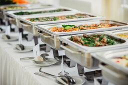 Aufgereihte Catering-Speisen auf einem Tisch mit weißer Tischdecke und silbernen Wärmebehältern