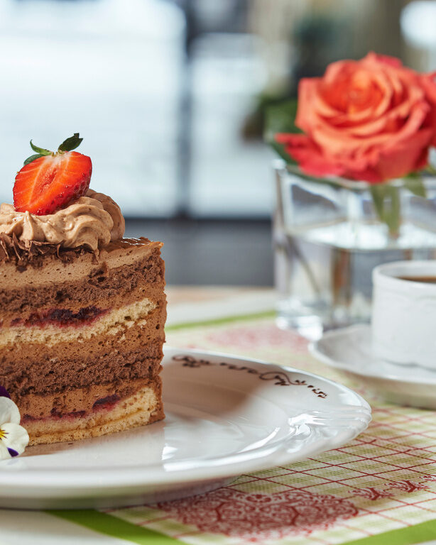 Tortenstück mit Erdbeere auf Teller, dahinter Vase mit Rose und Kaffeetasse