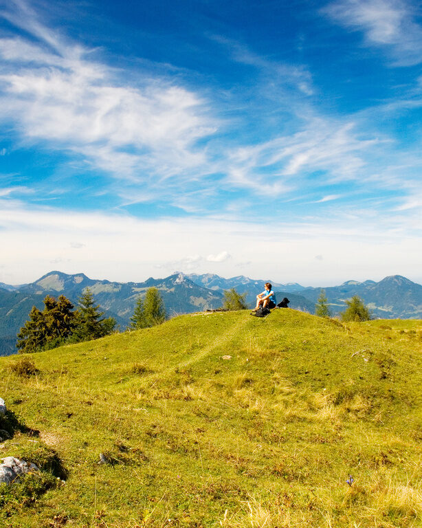 Frau sitzt auf Wiese auf Berg an sonnigem Tag, dahinter Alpen-Panorama und klarer Himmel