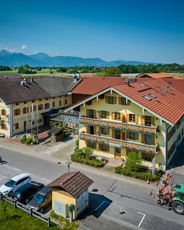 Luftaufnahme Vorderansicht Dreiseithof an sonnigem Tag, davor Parkplatz, dahinter Blick auf Alpen