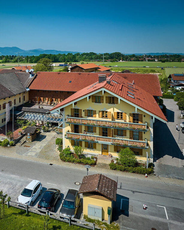 Luftaufnahme Vorderansicht Dreiseithof an sonnigem Tag, davor Parkplatz, dahinter Blick auf Alpen