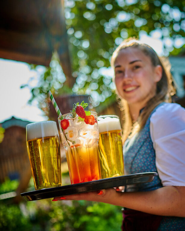 Eine gut gelaunte Bedienung trägt ein Tablet mit Getränken durch den Biergarten des Hotels Happinger Hof in Oberbayern.