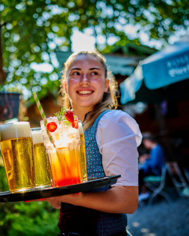Lächelnde Bedienung trägt Tablett mit Getränken an sonnigem Tag durch den Biergarten des Hotels