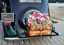 Ein schwarzer Oldtimer, der als Hochzeitsauto genutzt wird, hat zwei Koffer und Blumen am Heck