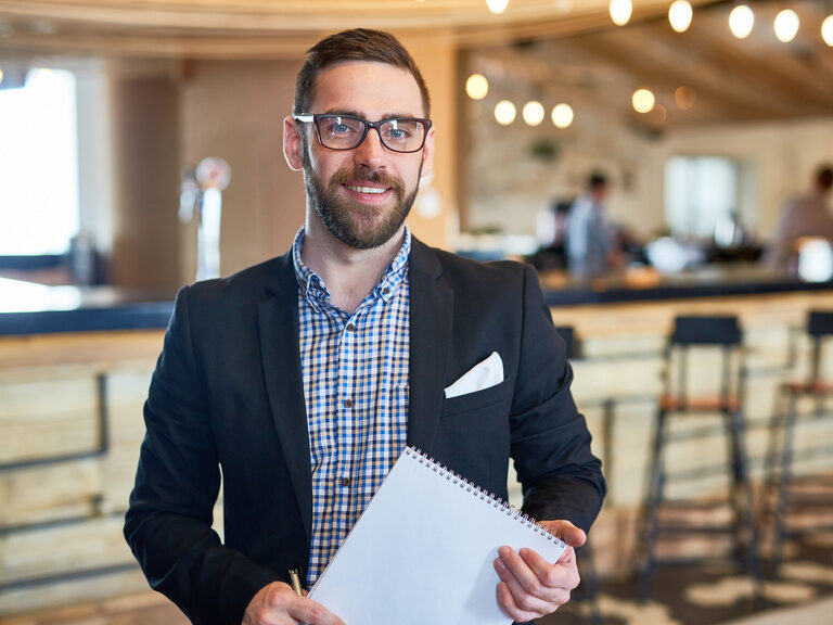 Ein Hotelfachmann im Hotel Happinger Hof in Rosenheim lächelt in die Kamera und hält einen Block in der Hand