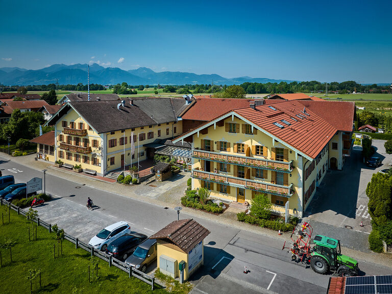 Luftaufnahme Vorderansicht Dreiseithof an sonnigem Tag, davor Parkplatz, dahinter Blick auf Alpen