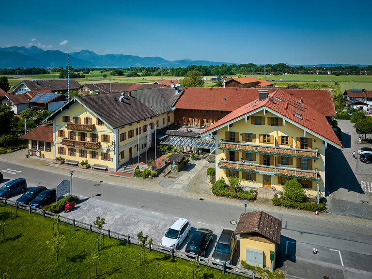 Luftaufnahme Vorderansicht Dreiseithof an sonnigem Tag, davor Parkplatz, dahinter Blick auf Alpen