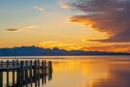 Ein malerischer Sonnenuntergang über der Wasseroberfläche des Chiemsees, es ist zudem ein Steg zu sehen.