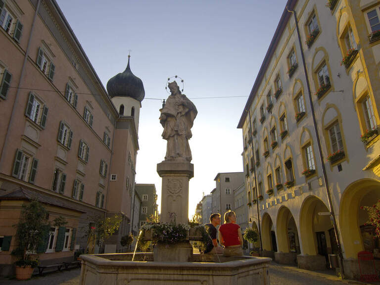 Springbrunnen in Altstadt Rosenheim nahe dem 3-Sterne-S-Hotel und Landgasthof Happinger Hof beim Chiemsee Springbrunnen aus Stein mit Kirchenfigur bei Dämmerung in oberbayerischer Altstadt